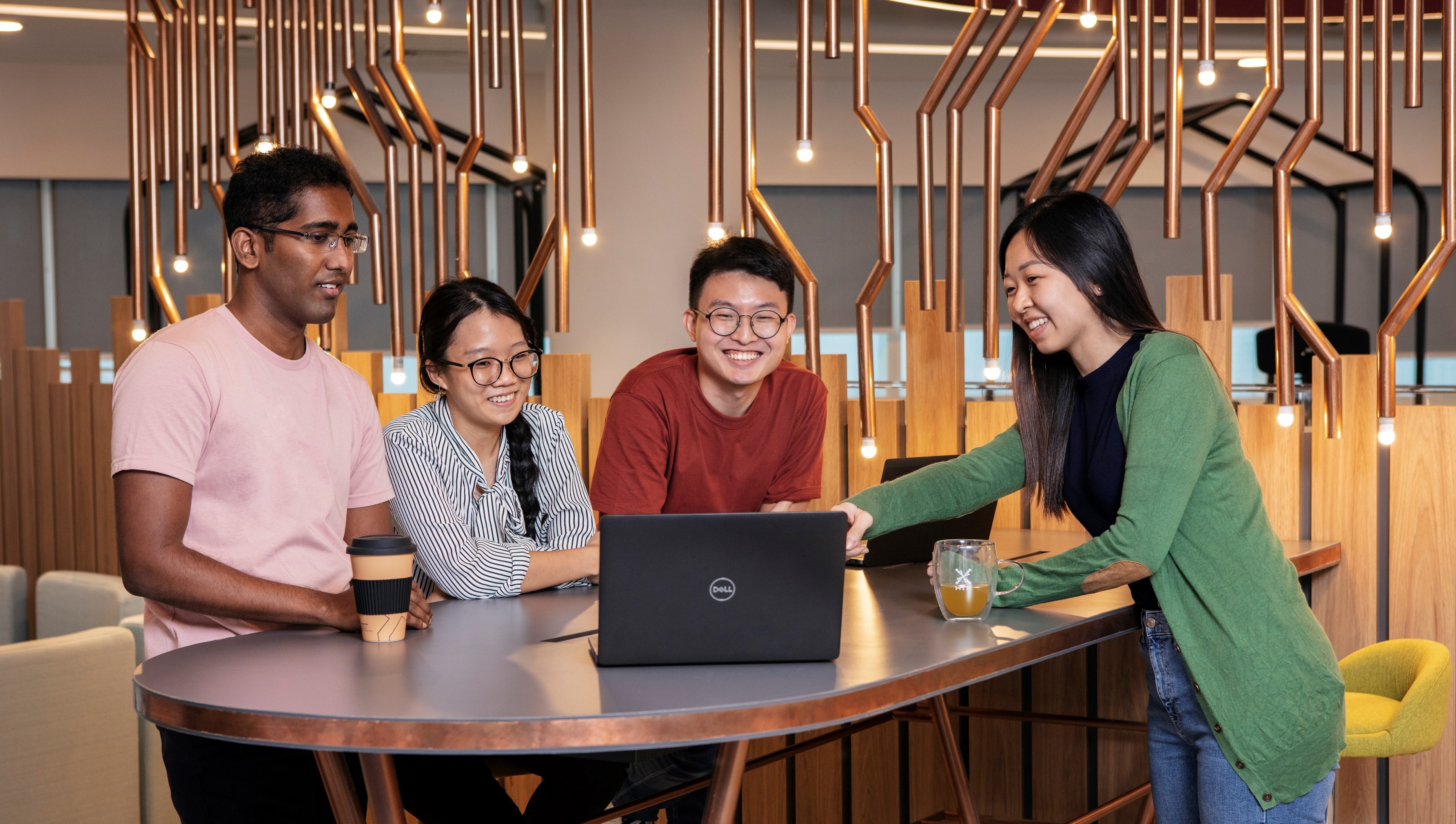 Four colleagues collaborating around a laptop in HTX's modern office space, smiling and engaged in discussion.